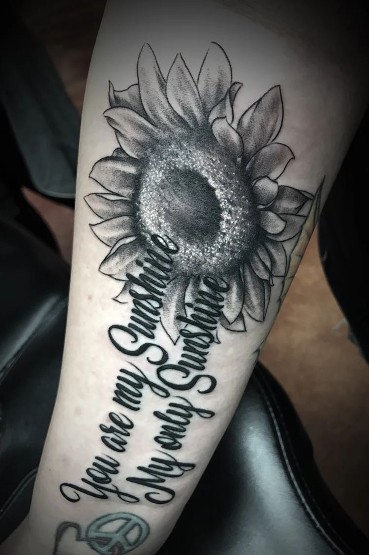 Black and grey sunflower with script lettering on the forearm